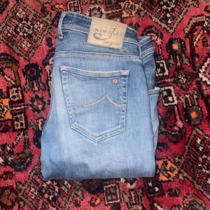 Blå jeans från Jacob Cohen - Snygga blå jeans från Jacob Cohen med klassisk femficksdesign och ljus tvätt. Byxorna har raka ben och diskreta broderade detaljer på fickan. Perfekta för en avslappnad och stilren look. de defekter som finns på varan syns på första och sista bilden  1(loggan) 5(2 hål i skrevet)