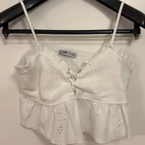 Vit croppad linnetopp från FBSister - Säljer en söt vit croppad linnetopp från FBSister med tunna axelband, volangdetaljer och broderade mönster. Toppen har en liten knytning framtill och är perfekt för varma dagar.