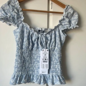 Ljusblå blus Gina Tricot - Supersöt ljusblå blus med smockad midja och volangdetaljer. Blusen har korta puffärmar, knytning fram och ett diskret mönster i vitt. Perfekt för vår och sommar! Aldrig använd 🌸