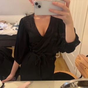 Svart omlottklänning med knytband - Säljer en svart omlottklänning med v-ringning och knytband i midjan. Klänningen har långa ärmar och en elegant, enkel design som passar perfekt till olika tillfällen.