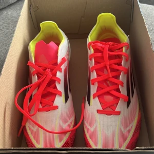 Adidas fotbollsskor med neonröda detaljer - Säljer ett par Adidas fotbollsskor i vitt med neonröda och gula detaljer. Skorna har snörning och en lätt, smidig design med mönstrad sula för bra grepp på planen. Perfekta för dig som vill sticka ut på fotbollsplanen! Dem är använda bara en gång och ser helt nya ut. Nyligen köpta och kan inte lämna tillbaks dem och skorna var stora för mig! Ganska snygga och fina skor. Pris kan diskuteras