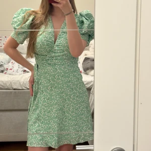 Grön klänning - Säljer denna super fina klänningen från adoore. Den är blommig med vita blommor. Man kan justera midjan så att den sitter hur man vill, vilket också gör det tajtare vid bysten. Aldrig använd utan bara testad, lappen kvar. Köpte för att ha på studentfirande men valde en annan, så hör av dig vid frågor🥰