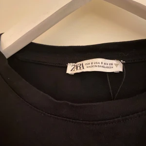 Svart volangtopp från Zara - Säljer en svart topp från Zara med korta volangärmar. Toppen har en rak passform och är tillverkad i ett mjukt material. Perfekt att styla med både jeans och kjol.