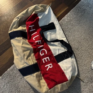 Beige och röd weekendbag från Tommy Hilfiger - Säljer en stor och rymlig weekendbag från Tommy Hilfiger. Den har svarta handtag och axelrem samt detaljer i marinblått. Perfekt för resor eller gymmet.