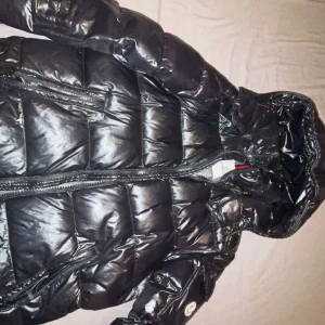 Moncler jacka - Säljer en svart pufferjacka från Moncler med glansig finish och huva. Jackan har dragkedja framtill, två sidofickor med dragkedja och Moncler-logga på ärmen. Insidan har ett unikt serietidningsmönster. Perfekt för kalla dagar.