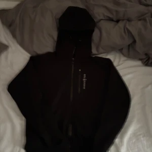 Svart sailracing hoodie  - Svart hoodie från Sail Racing med dragkedja framtill och logga på bröstet. Jackan har huva och en ficka med dragkedja. Perfekt för en sportig och avslappnad stil.