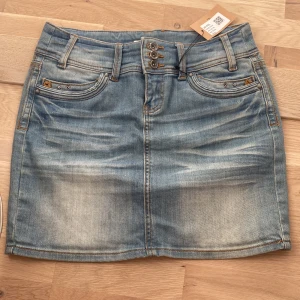 Blå jeanskjol med dubbla knappar - Snygg blå jeanskjol med slitningar och dubbla knappar framtill. Klassisk femficksmodell med bälteshällor och kontrastsömmar. Perfekt för en avslappnad och trendig look.
