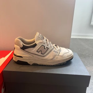 Vita New Balance 550 sneakers - Säljer ett par klassiska off-vita New Balance 550 sneakers med svarta detaljer och snörning. Skorna har en rund tå och platt sula, perfekt för en sportig och stilren look. Ikonisk N-logga på sidan och bekväm passform.