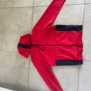 Röd hoodie med svarta detaljer från Hugo Boss - Röd hoodie med svarta paneler på sidorna och en svart rand på huvan. Dragkedja framtill och justerbar huva med snören. Boss-logga på bröstet. Perfekt för en sportig och avslappnad stil.