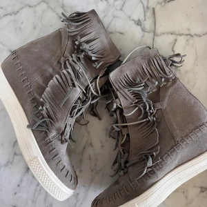 Grå mockaboots med fransar - Säljer ett par grå boots i mocka med högt skaft och coola fransdetaljer runt hela skon. Skorna har snörning framtill och en vit platt sula som ger en snygg kontrast. Perfekta för dig som vill sticka ut med din stil! Kan sälja för billigare vi smidig affär