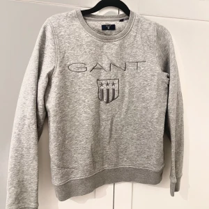 Grå sweatshirt från GANT - Säljer en klassisk grå sweatshirt från GANT med broderad logga och sköld framtill. Tröjan har rund halsringning och långa ärmar. Perfekt för en avslappnad och stilren look.