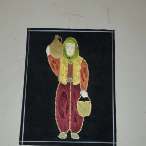 Broderad tavla med traditionell klädsel - Broderad bild på en person i traditionella byxor med vida ben i vinröd färg och gulddetaljer. Byxorna har en lös passform och är dekorerade med broderier. Motivet visar även en väst och sjal i grönt och gult. Det är 100% handgjord. Kontakta för mer information.