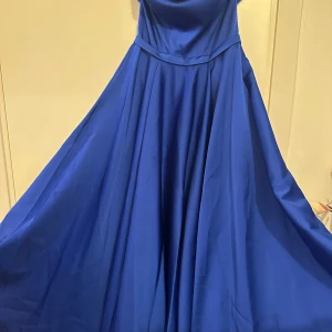 Blå långklänning med glittrig detalj - Säljer en elegant blå långklänning med axelbandslös design och glittriga pärldetaljer längs överdelen. Klänningen har en vacker, svepande kjol och är perfekt för bal eller fest. Materialet är glansigt och ger en lyxig känsla.