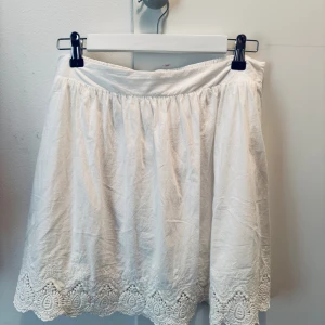 Vit kjol med spetskant från ONLY - Söt vit kjol från ONLY med bred resår i midjan och vacker broderad spetskant nertill. Kjolen är kort och har en luftig passform, perfekt för varma dagar. Tillverkad i bomull för en skön känsla. Storlek 36