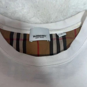 Vit t-shirt med rutiga ärmar från Burberry - Säljer en vit t-shirt från Burberry med klassiska beige, svarta och röda rutiga ärmar. T-shirten har rund halsringning och är tillverkad i bomull. Perfekt för dig som gillar stilrena plagg med en twist. Lite oversized, tvär fet men lite liten för mig. Snabb affär kan säljas för snabbare. 