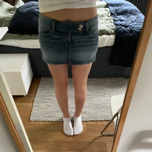 Blå jeanskjol med knapp och fickor - Säljer en klassisk blå jeanskjol med knapp och dragkedja framtill. Kjolen har två fickor fram och två bak, samt en kort passform som ger en snygg och avslappnad look.