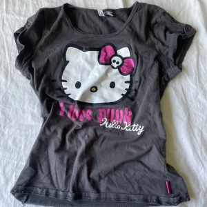 Hello Kitty punk t-shirt - Svart t-shirt med stort Hello Kitty-tryck på framsidan, där Hello Kitty har en rosa rosett med dödskalle. Texten 'I love punk' i rosa och vitt. Perfekt för dig som gillar en edgy stil! Köp nu går bra eller skriv om du har frågor💗