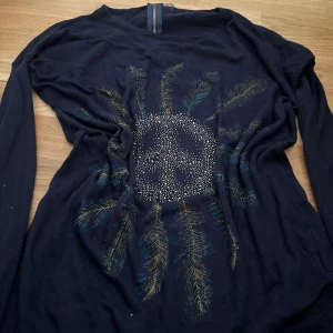 Mörkblå långärmad tröja med paljettmönster - Säljer en mörkblå långärmad tröja med dekorativt paljettmönster framtill som liknar fjädrar runt en cirkel. Tröjan har dragkedja i nacken och ribbade ärmar. Perfekt för dig som vill sticka ut lite extra.