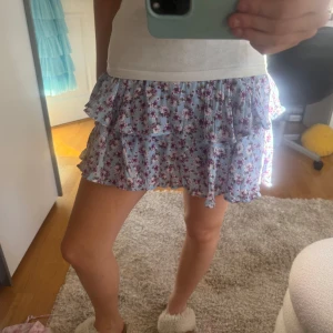 Blommig volangkjol i ljusblått - Supersöt kort kjol i ljusblått med blommigt mönster och volanger. Perfekt för varma dagar och ger en romantisk vibe. Resår i midjan för skön passform.