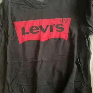 Svart t-shirt från Levi's med röd logga - Svart t-shirt från Levi's med den klassiska röda loggan tryckt framtill. T-shirten har rund halsringning och är tillverkad i mjuk bomull. Perfekt för en avslappnad och stilren look.