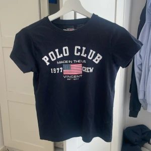 Mörkblå t-shirt från Polo Club - Mörkblå t-shirt från Polo Club Saint Vincent med vit text och amerikansk flagga tryckt på bröstet. Klassisk rund halsringning och korta ärmar. Perfekt för en avslappnad och sportig stil.