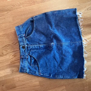 Blå jeanskjol med rå kant - Säljer en klassisk blå jeanskjol med rå, fransig kant nertill och röda detaljer vid fickor och bälteshällor. Kjolen har normal passform och är kort i modellen. Perfekt att styla med en enkel topp eller hoodie.