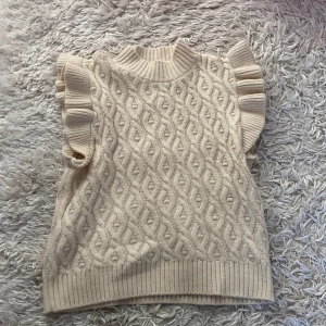 Beige stickad ärmlös tröja med volang - Superfin beige stickad tröja med höghals och volangdetaljer vid axlarna. Den är i storlek 146-152 men är nästan lite för stor för mig som i vanliga fall har 158-164! Endast använd 1 gång!!