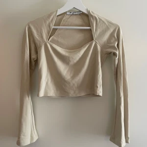 Beige croppad långärmad topp från NA-KD - Trendig beige croppad topp från NA-KD med lång ärm och unik halsringning. Tillverkad i mjuk bomullsmix som sitter skönt mot kroppen. Aldrig använt.