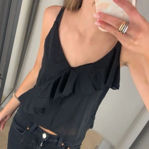 Svart linnetopp med volang - Säljer en svart linnetopp med smala axelband och snygg volangdetalj framtill. Toppen har v-ringning och är tillverkad i ett lätt material som passar perfekt till sommaren.