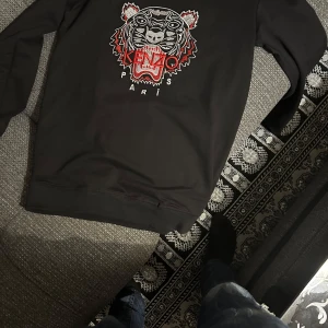 Svart sweatshirt med tigerbrodyr från Kenzo - Svart sweatshirt från Kenzo med ikonisk tigerbrodyr och röd Kenzo-logga framtill. Tröjan har rund hals och långa ärmar. Perfekt statement-plagg för dig som gillar streetwear och snygga detaljer. Priset går att diskutera om så önskas 