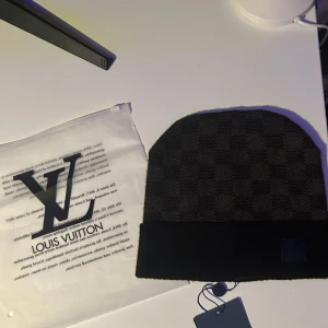 Svart Rutig mössa - Snygg och exklusiv mössa i svart och grått med diskret rutigt mönster. Mössan har en ribbad kant och en svart läderpatch med L-v-logga framtill. Perfekt accessoar för kallare dagar.