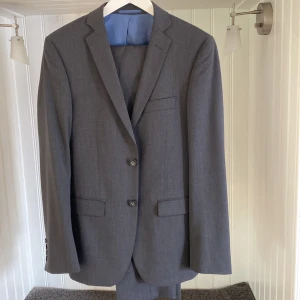 Grå kostym från Dressmann Slim Fit - Säljer en stilren grå kostym från Dressmann i slim fit-modell. Kavajen har två knappar, klassiska fickor och matchande byxor. Perfekt för bal eller fest.