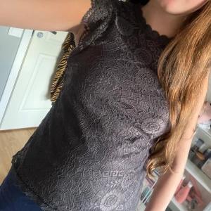 Grå spetsblus med volangärmar - Säljer en grå blus i spets med fina blommönster och volangdetaljer längs ärmarna. Blusen är ärmlös och har en liten knapp i nacken. Perfekt för dig som gillar romantiska detaljer och vill ha något extra till outfiten.