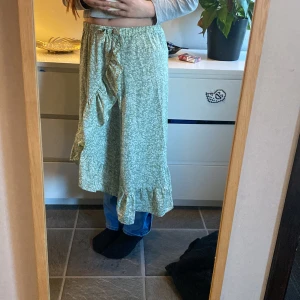 Grön blommig omlottkjol med volang - Säljer en ljusgrön omlottkjol med småblommigt mönster och volangkant nertill. Kjolen har knytband i midjan och är i en luftig modell som passar perfekt till varmare dagar. Stretch i midjan