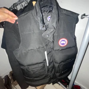Svart dunväst från Canada Goose - Säljer en svart dunväst från Canada Goose med klassisk logotyp på bröstet. Västen har hög krage, knappar och dragkedja framtill samt två stora fickor. Perfekt för lager-på-lager och håller dig varm under kyliga dagar.