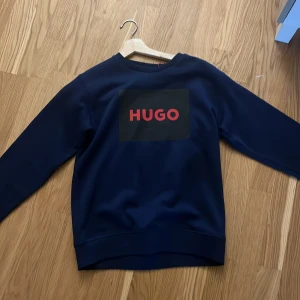 Mörkblå sweatshirt från Hugo - Säljer en mörkblå sweatshirt från Hugo med stor röd logga framtill. Tröjan har rund halsringning och långa ärmar. Perfekt för en stilren och avslappnad look.