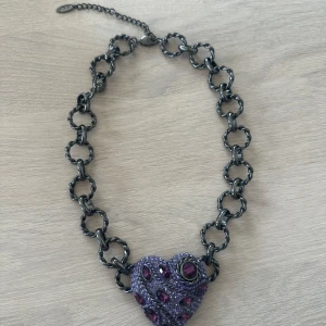 Kedjehalsband med lila hjärthänge - Säljer ett chunky halsband med stora, mörka metallringar och ett stort hjärthänge dekorerat med lila och mörklila stenar. Hängsmycket har glittriga detaljer och en unik, iögonfallande design. Perfekt för dig som vill sticka ut!