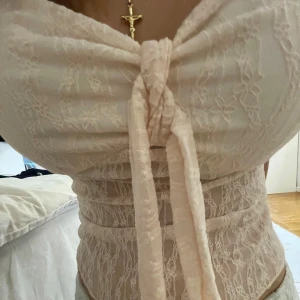 Ljusrosa spetsig topp - Säljer en ljusrosa topp i spets med knyt framtill. Toppen är axelbandslös och har en romantisk känsla med sitt transparenta spetsmaterial och fina detaljer. Perfekt för att skapa en feminin look.