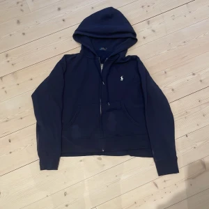 Mörkblå hoodie från Polo Ralph Lauren - Säljer en klassisk mörkblå hoodie från Polo Ralph Lauren med dragkedja och huva. Tröjan har två fickor framtill och den ikoniska loggan broderad på bröstet. Perfekt för en avslappnad stil. Skick 7/10, det är bara att skicka om ni undrar något!