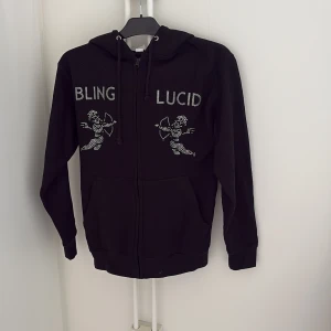 Svart hoodie med strasstenar från Bling Lucid - Svart hoodie från Bling Lucid med dragkedja och huva. Framsidan har texten 'BLING LUCID' och två änglar i strasstenar. Tillverkad i bomull och polyester. Perfekt för dig som gillar unika detaljer.