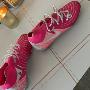 Nike fotbollsskor i rosa och vitt - Säljer ett par Nike fotbollsskor i en snygg rosa färg med vita detaljer och snörning. Skorna har en högre strumpliknande krage och mönstrad ovansida för bättre bollkontroll. Sulan är silvrig med dobbar för spel på gräs. Används en gång . Och säljs på grund av att det inte passade