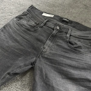 Gråa jeansbyxor från Replay - Säljer ett par svarta Replay jeans med klassisk femficksdesign och diskreta slitningar. Byxorna har normal passform och raka ben. Märkeslogga på bakfickan och läderpatch i midjan. Perfekta för en stilren look.