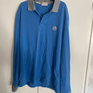 Moncler tröja - Säljer en blå långärmad pikétröja från Moncler storlek M med grå krage och muddar. Tröjan har klassisk pikéknäppning och Moncler-logga på bröstet. Perfekt för en sportig och stilren look. Nyskick knappt använd.