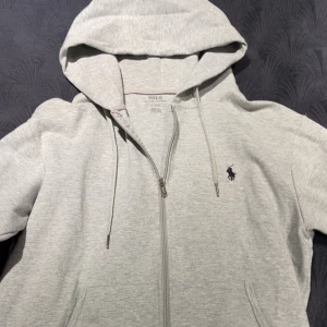 Polo hoodie - Säljer en stilren ljusgrå hoodie från Polo Ralph Lauren med dragkedja och klassisk logga broderad på bröstet. Tröjan har huva med dragsko och två fickor framtill. Perfekt för en avslappnad look. Ny pris 1800