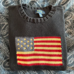 Ralph Lauren tröja - Säljer den virala mörkblå stickad tröja från Polo Ralph Lauren med en stor amerikansk flagga på bröstet. Tröjan har en unisex passform i storlek 14-16 år men skulle säga att den sitter som en S/M vuxen storlek. Tröjan ser grå ut på bilden men är mörkblå i verkligheten!