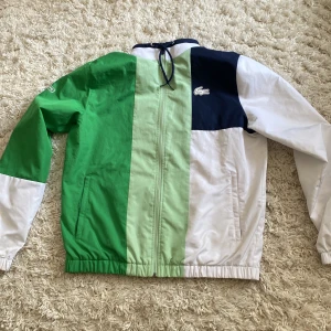 LacosteMedvedev Träningsoverall Herrar - Säljer en snygg vindjacka från Lacoste i blockfärger: grön, ljusgrön, vit och marinblå. Jackan har dragkedja, hög krage med snörning och elastiska muddar. Klassisk Lacoste-logga på bröstet. Perfekt för dig som gillar sportig stil. Tröjan säljer jag för 399 men om man vill köpa med byxorna kan du köppa för 599 tillsammans. Byxorna är lite sönder som man kan se på bild 5 och ett litet märke på den också.  Annars är den super bra. Köpte nytt för 1800. Pris kan diskuteras. Skriv för mer information,