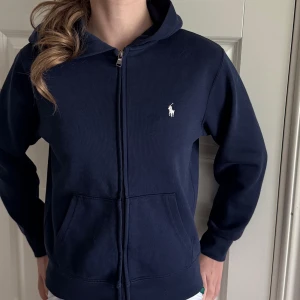 Mörkblå hoodie med dragkedja från Ralph Lauren - Mörkblå hoodie från Ralph Lauren med dragkedja framtill och klassisk vit logga på bröstet. Tröjan har huva och två fickor framtill. Perfekt för en avslappnad stil. Motsvarar ca stl 152 eller ålder 10-12 år