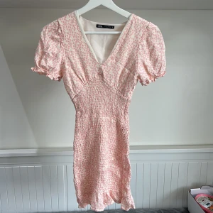 Rosa blommig kortklänning från Zara - Supersöt kortklänning från Zara i ljusrosa med små blommor. Klänningen har v-ringning, puffärmar och smockad midja som ger en fin passform. Perfekt för sommarens alla tillfällen!