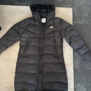 Svart lång dunjacka från Nike - Säljer en svart lång dunjacka från Nike med huva och dragkedja. Jackan har Storm-FIT-teknologi som skyddar mot väder och vind. Perfekt för kyliga dagar och har en stilren design med Nike-logga på bröstet.