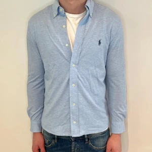Ljusblå skjorta från Ralph Lauren - Säljer en klassisk ljusblå skjorta från Ralph Lauren med button-down krage och broderad logga på bröstet. Skjortan har långa ärmar och vita knappar framtill. Perfekt för en stilren look.